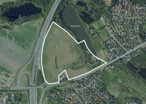 Luftfotoet af et grønt areal afgrænset med hvid streg, omgivet af motorvej, rundkørsel, søen Vestermose og boligområder.